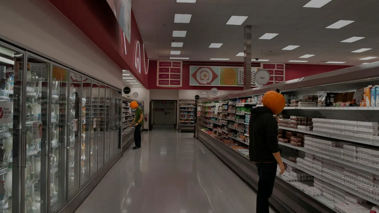 Two people in a grocery store aisle wearing pumpkins on their heads. One browses refrigerated goods; the other examines dairy products.