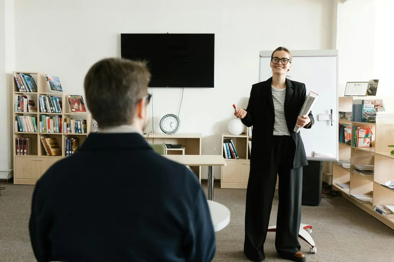 A woman in business attire stands smiling, holding a folder and pen in a library setting. She faces a seated person. Bookshelves and a whiteboard surround them.