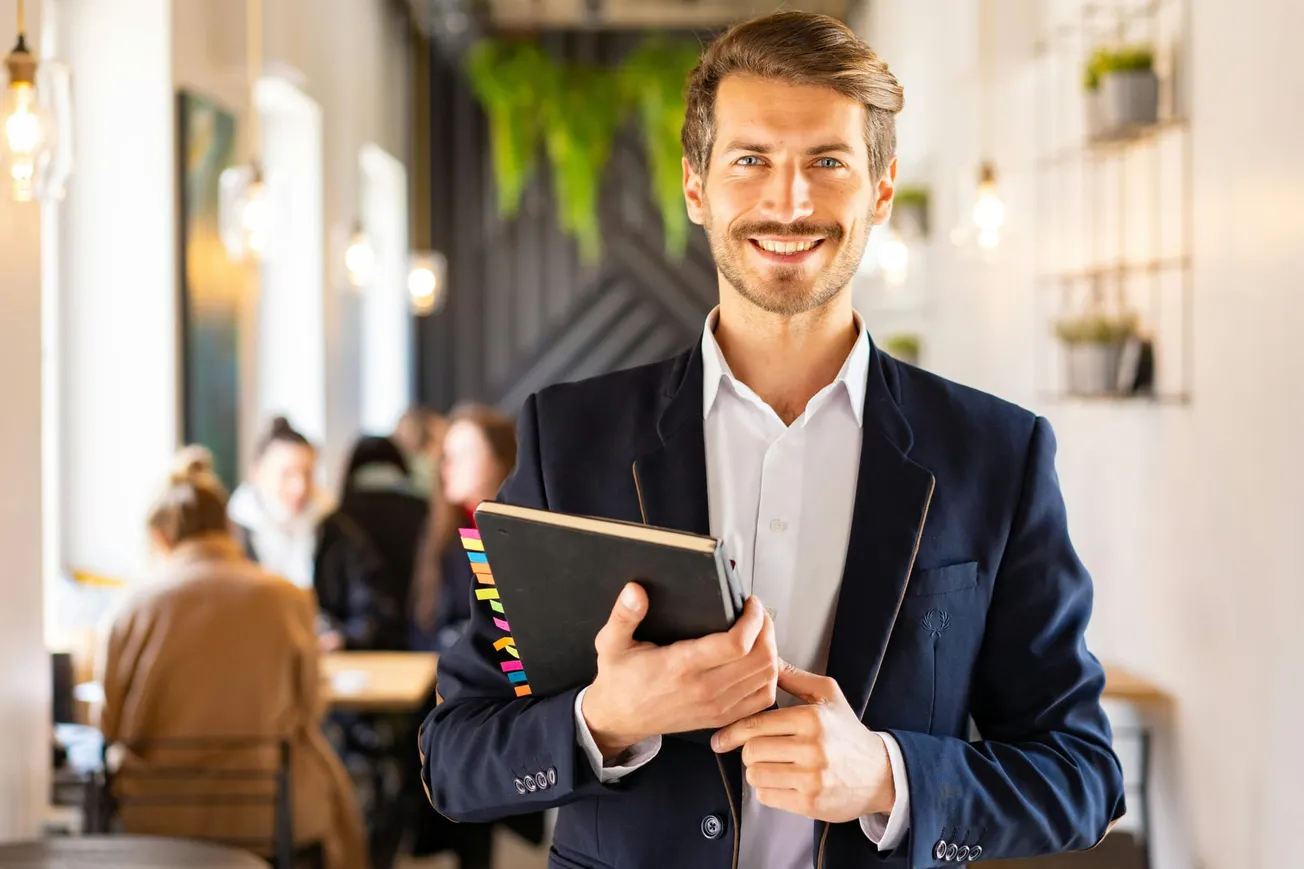 Smiling man in a suit holds a notebook in a cozy cafe. Soft lighting, blurred people in the background, and green plants create a welcoming ambiance.