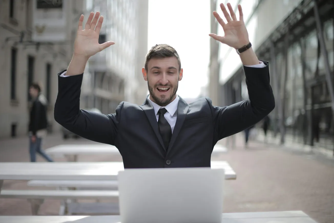 A man in a suit sits outside at a white table, looking at a laptop with raised hands and a joyful expression, against a blurred urban backdrop.