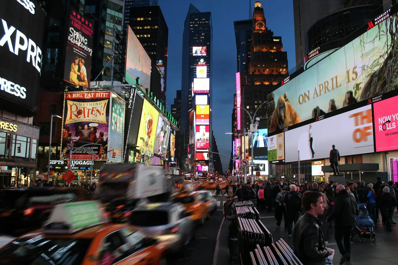 Bustling Times Square at dusk; vibrant billboards illuminate crowded streets with bright lights, taxis, and pedestrians in a lively, urban scene.