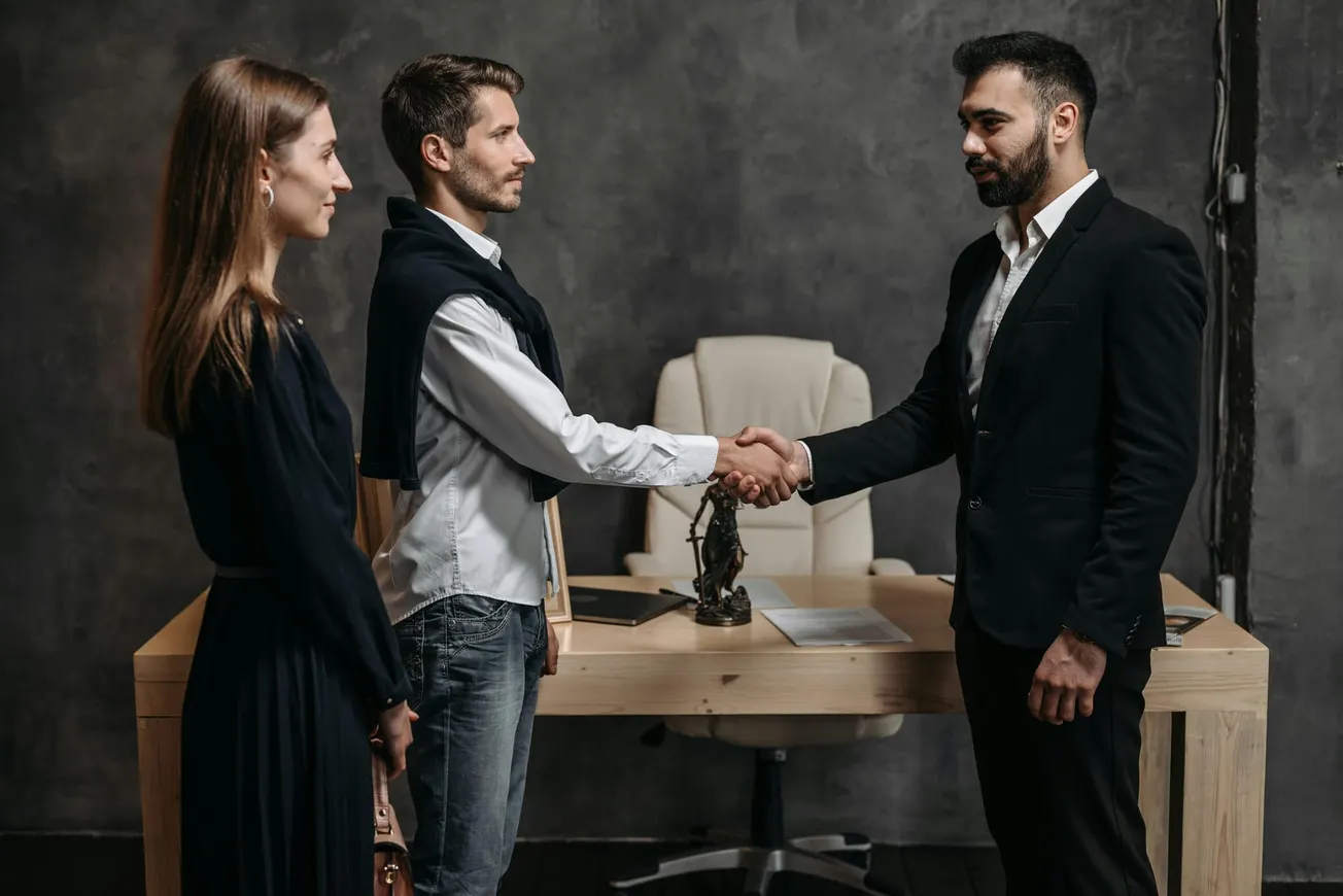 Two men shake hands in a dimly lit office, exuding professionalism and agreement, while a woman stands nearby observing. A desk with documents is behind them.