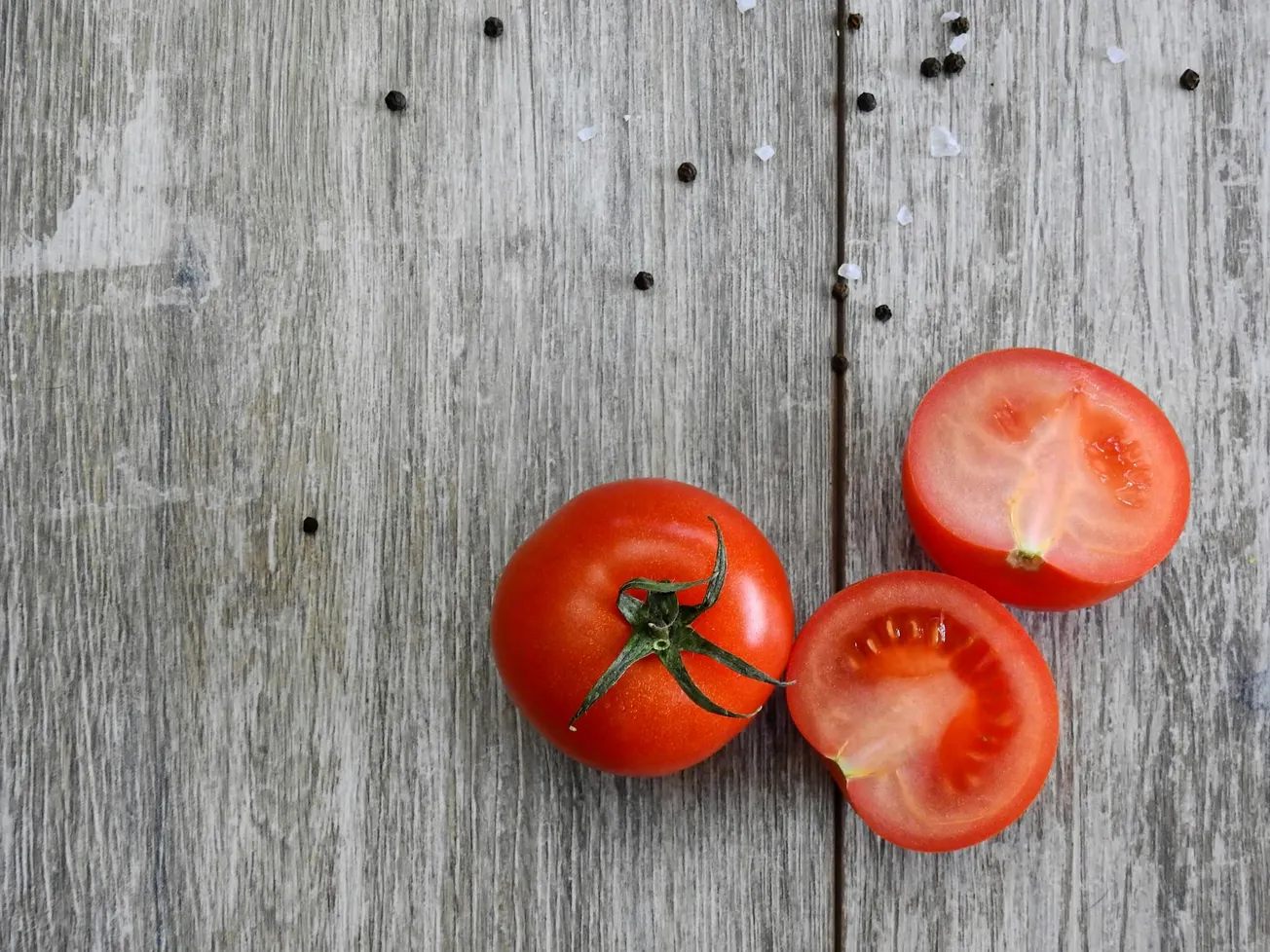 Three ripe tomatoes, one whole and two halved, rest on a rustic wooden surface. Scattered salt and pepper add texture to the minimalist scene.