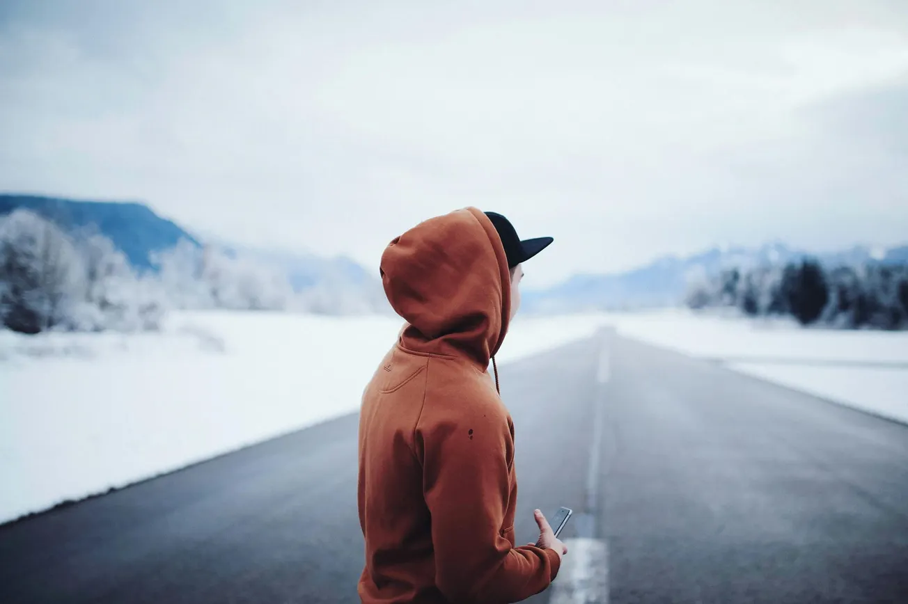 A person in an orange hoodie and cap stands on a deserted snowy road, holding a phone. Snow-covered trees and distant mountains create a calm, wintry scene.