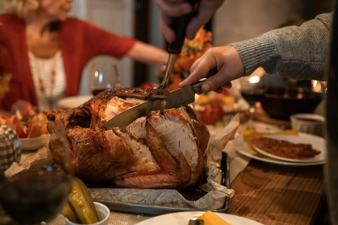Person slicing a roasted turkey at a festive dinner table, surrounded by various dishes. Warm, cozy atmosphere with family gathered in the background.