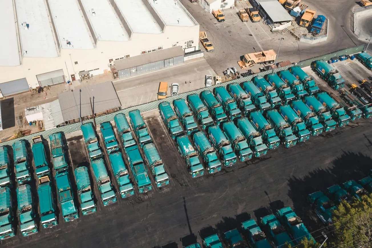 Aerial view of a large fleet of parked teal garbage trucks beside a beige industrial building. The scene conveys organization and efficiency.