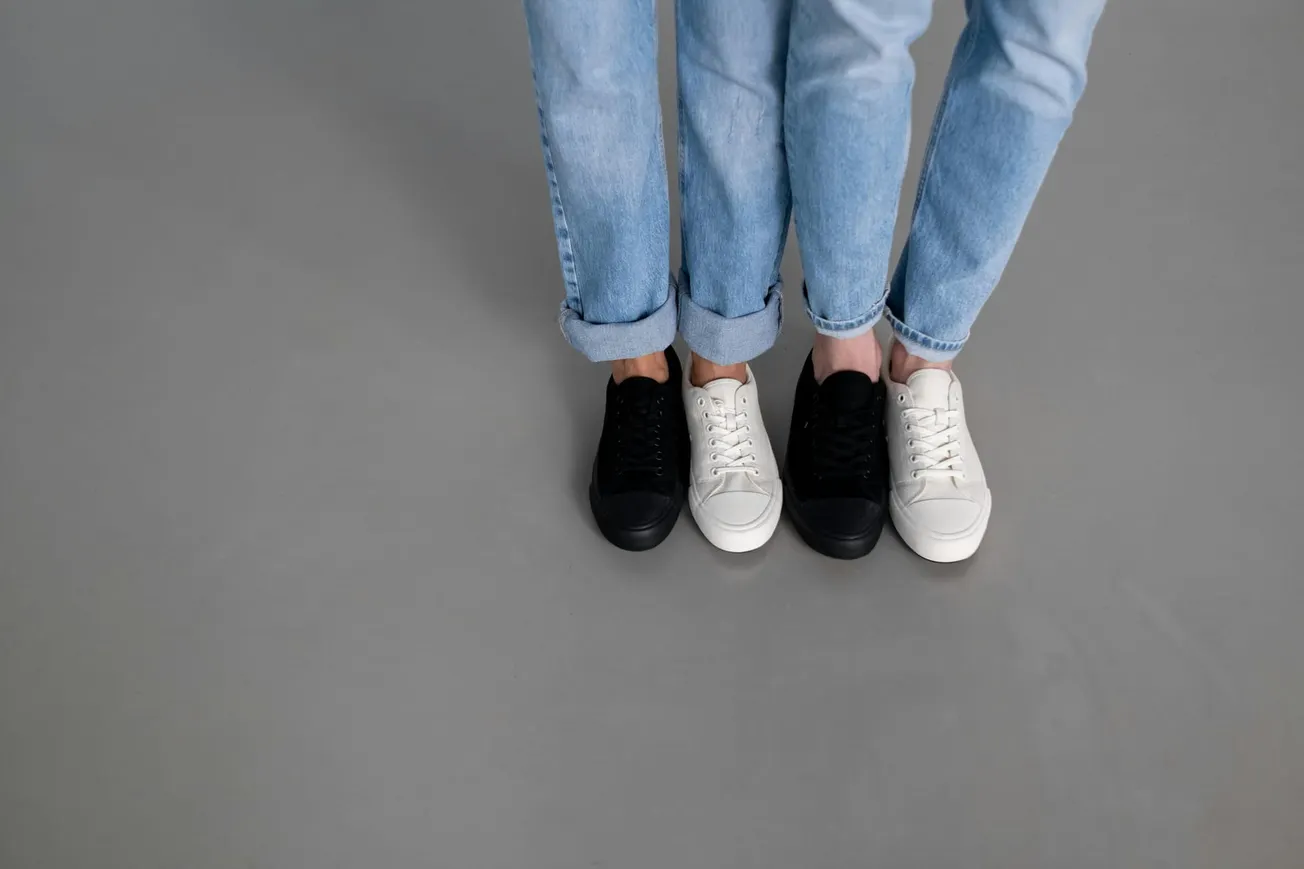Two people stand side by side on a grey floor, wearing rolled-up blue jeans. One wears black sneakers, the other mismatched black and white sneakers.