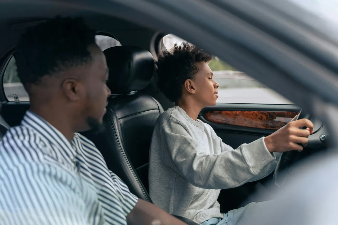 A person wearing a gray sweatshirt focuses on driving a car, while another person in a striped shirt calmly observes from the passenger seat.