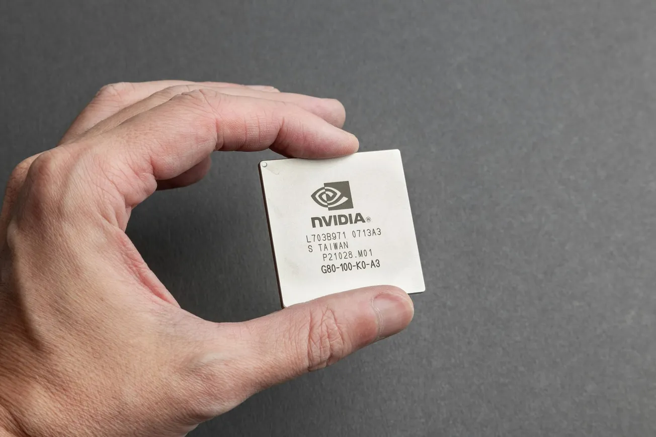 A hand holding an NVIDIA computer chip against a gray background. The chip is square, metallic, and engraved with technical specifications.