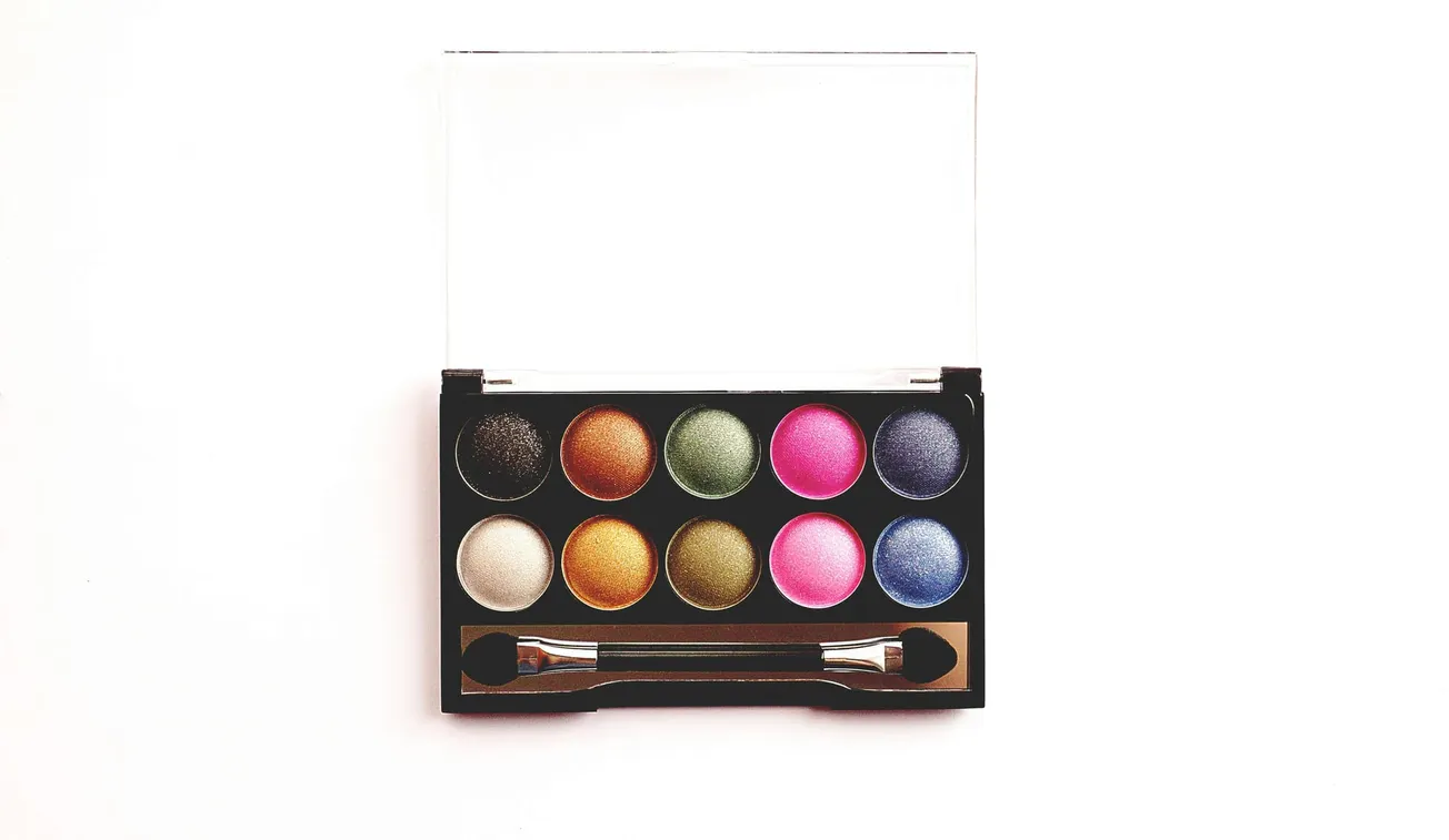 A makeup palette featuring ten circular eyeshadows in various colors, including black, green, pink, and blue, with an applicator below them.