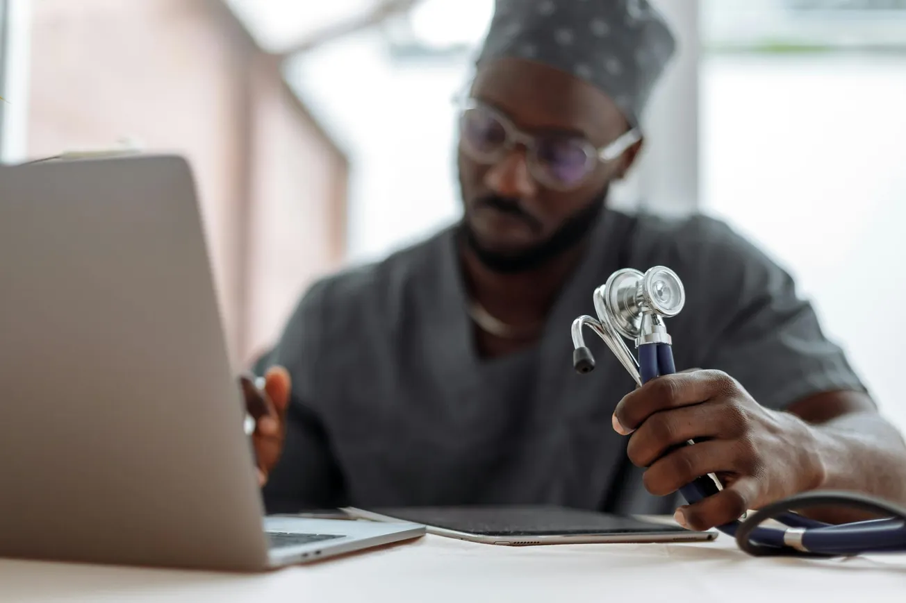 A doctor in scrubs holds a stethoscope while using a laptop in a bright room. The scene conveys focus and professionalism.