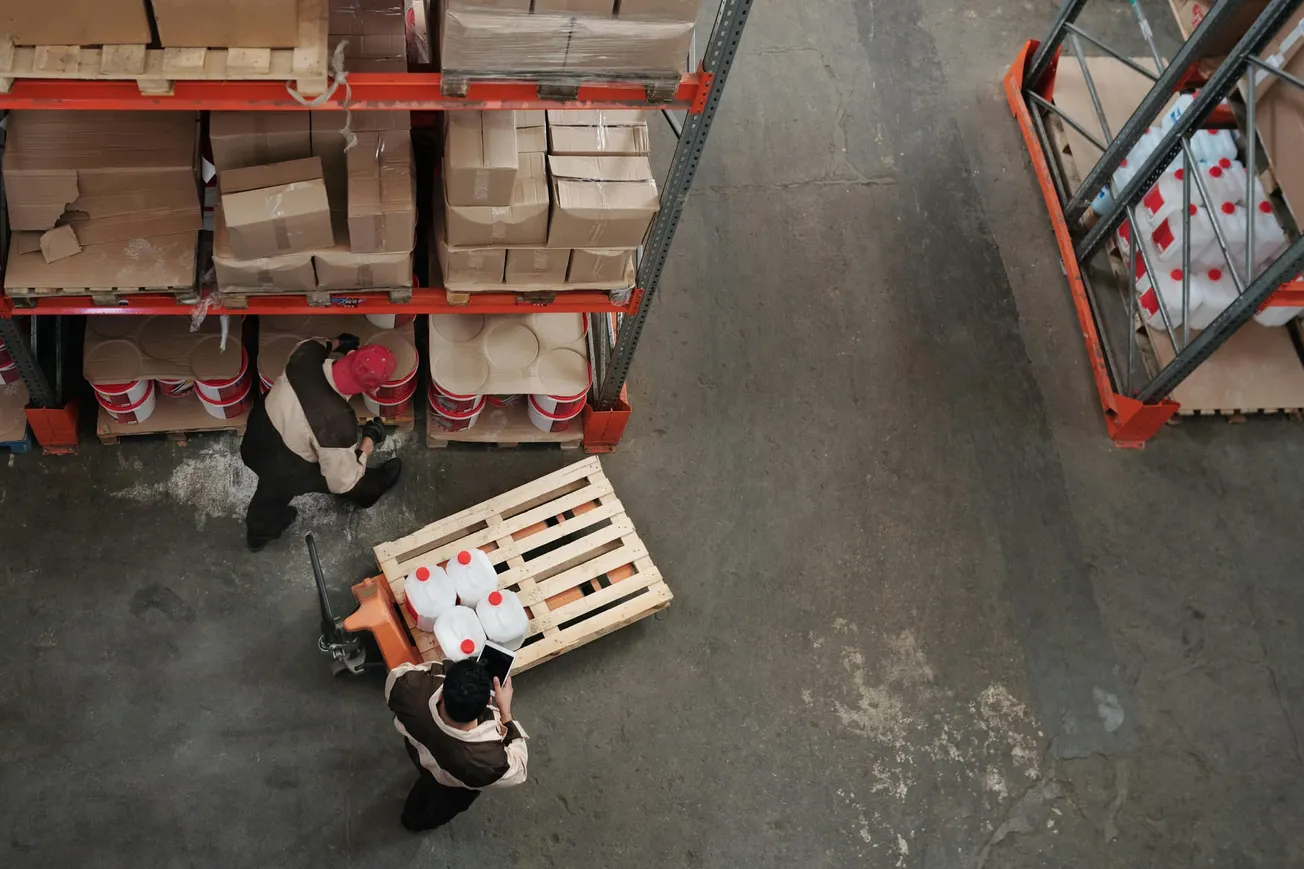 Two workers in a warehouse move large white containers on a pallet jack, surrounded by shelves filled with cardboard boxes and red-lidded buckets.