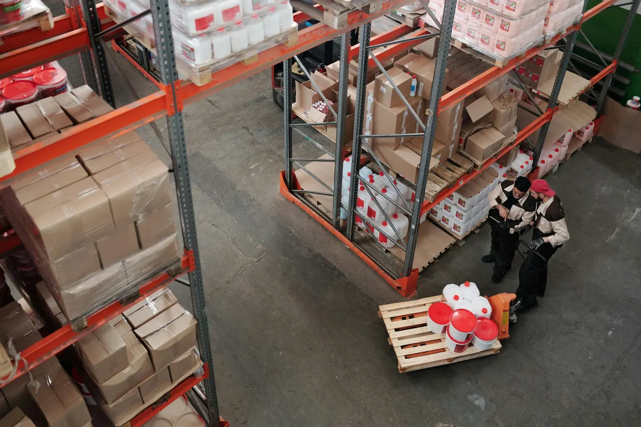 Warehouse interior from above, two workers in uniforms handle a pallet jack with white and red containers. Shelves stocked with boxes and containers surround them.