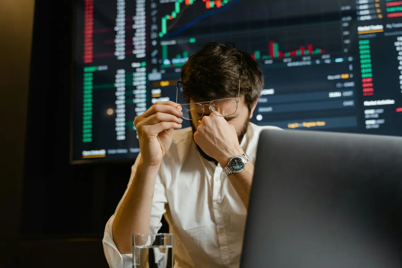 Man in a white shirt sits at a desk, holding glasses, looking stressed. In the background, a screen shows fluctuating stock market graphs.