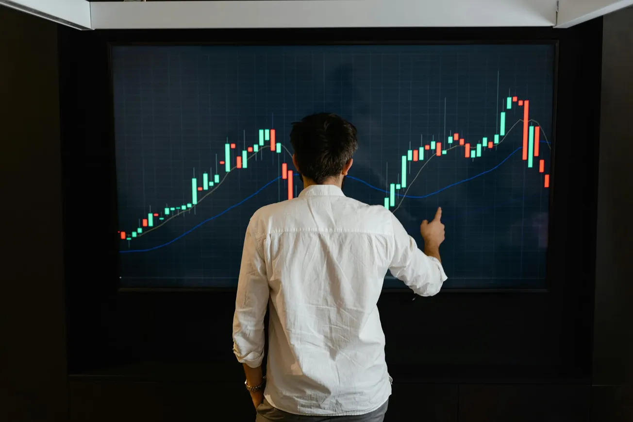 A person in a white shirt points to a digital screen showing a fluctuating stock market chart, conveying focus and analysis.