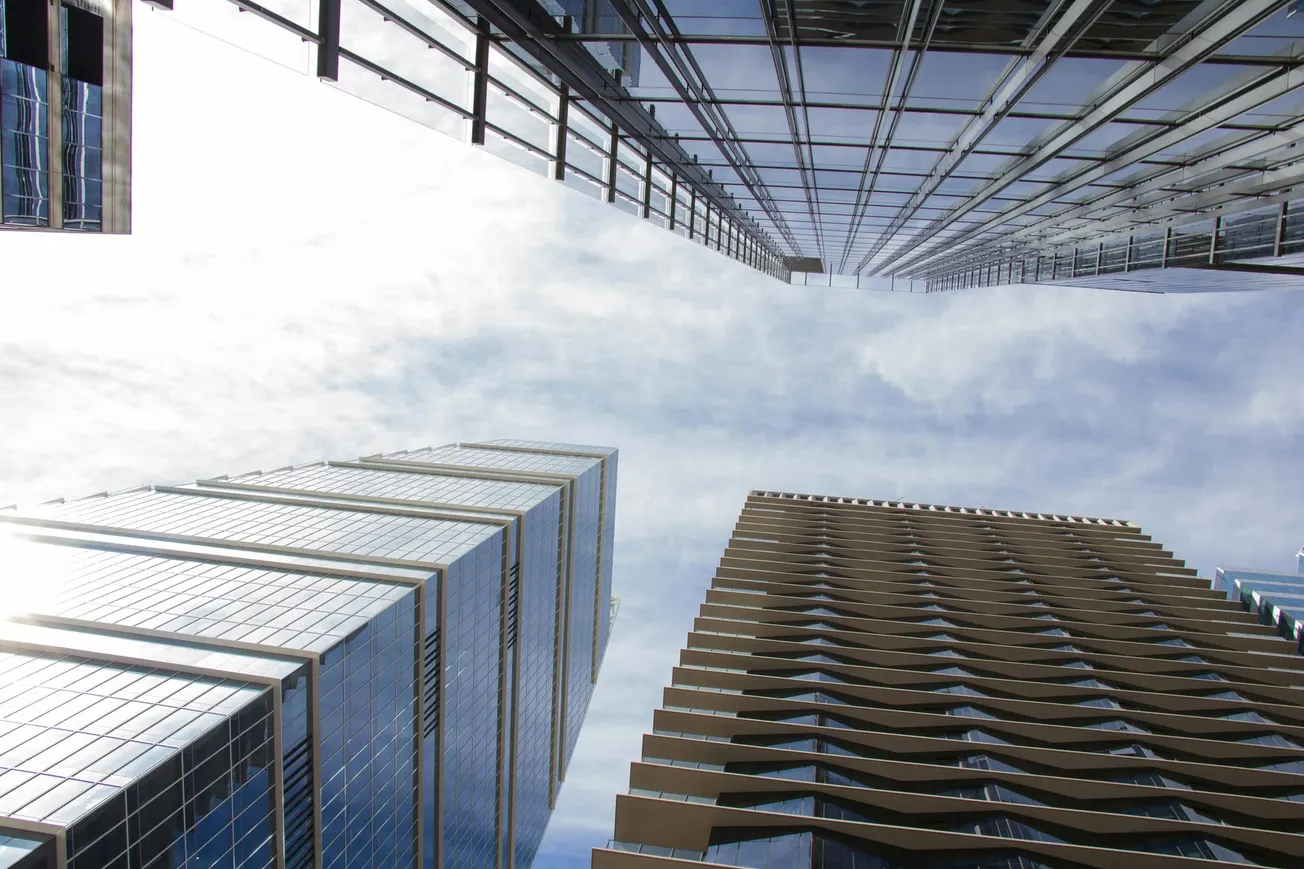 Looking up at three high-rise buildings with reflective glass and varied designs against a partly cloudy sky, conveying an urban and modern feel.