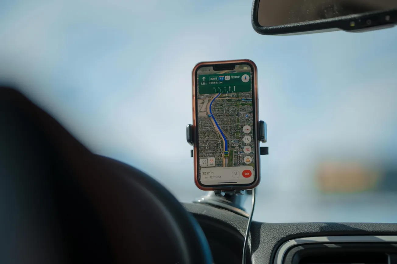 Smartphone with GPS map app in car mount shows navigation on a sunny day. Blue route, clear sky visible through windshield, suggesting travel.