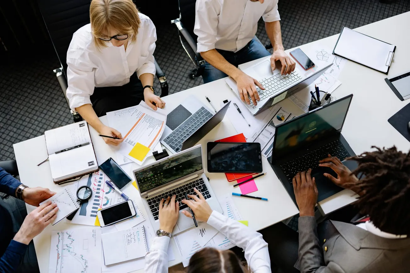 A diverse group of individuals collaborating at a cluttered desk with laptops, tablets, and papers, conveying a busy, productive office atmosphere.