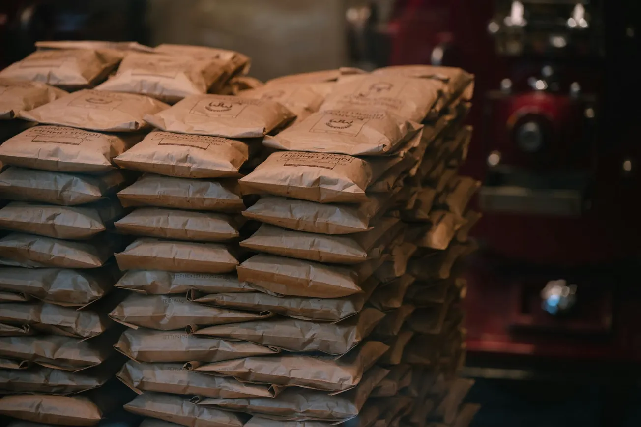 A stack of neatly arranged brown paper bags, likely containing coffee beans, sits beside a red industrial coffee roaster, creating a warm, artisanal atmosphere.