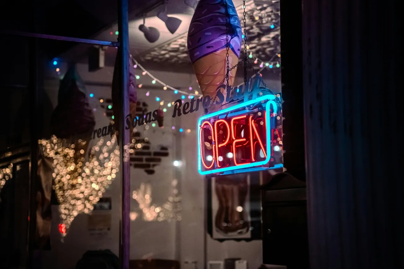A neon "OPEN" sign glows in red and blue outside a dimly lit storefront, with a large ice cream cone decoration and colorful string lights inside.