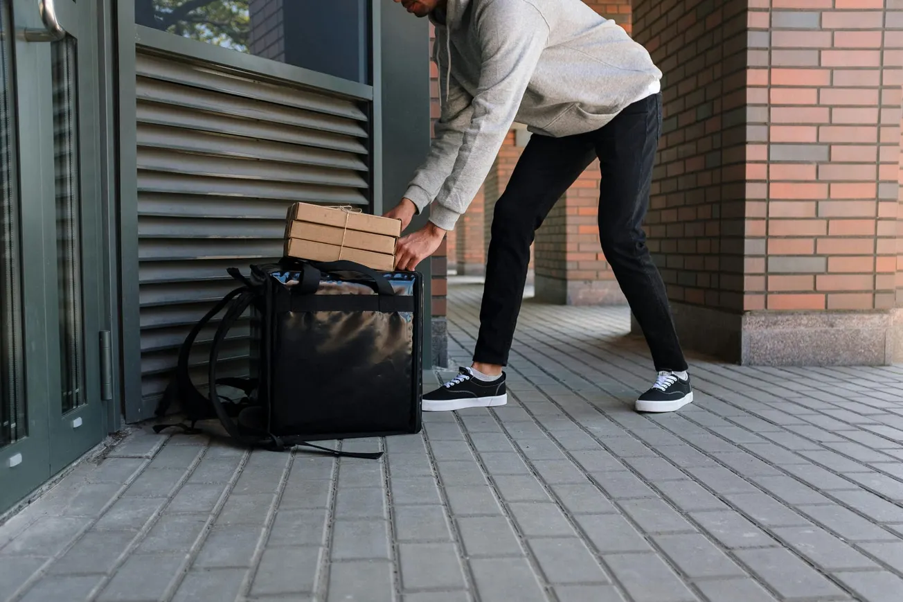A person in black pants and sneakers places two pizza boxes in a black delivery bag on a brick sidewalk. The setting is urban and appears calm.