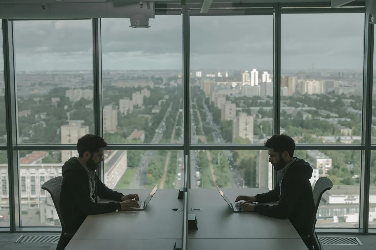 Two identical individuals sit at a table in a modern office, back-to-back, using laptops. A large window showcases a cityscape with a central road.