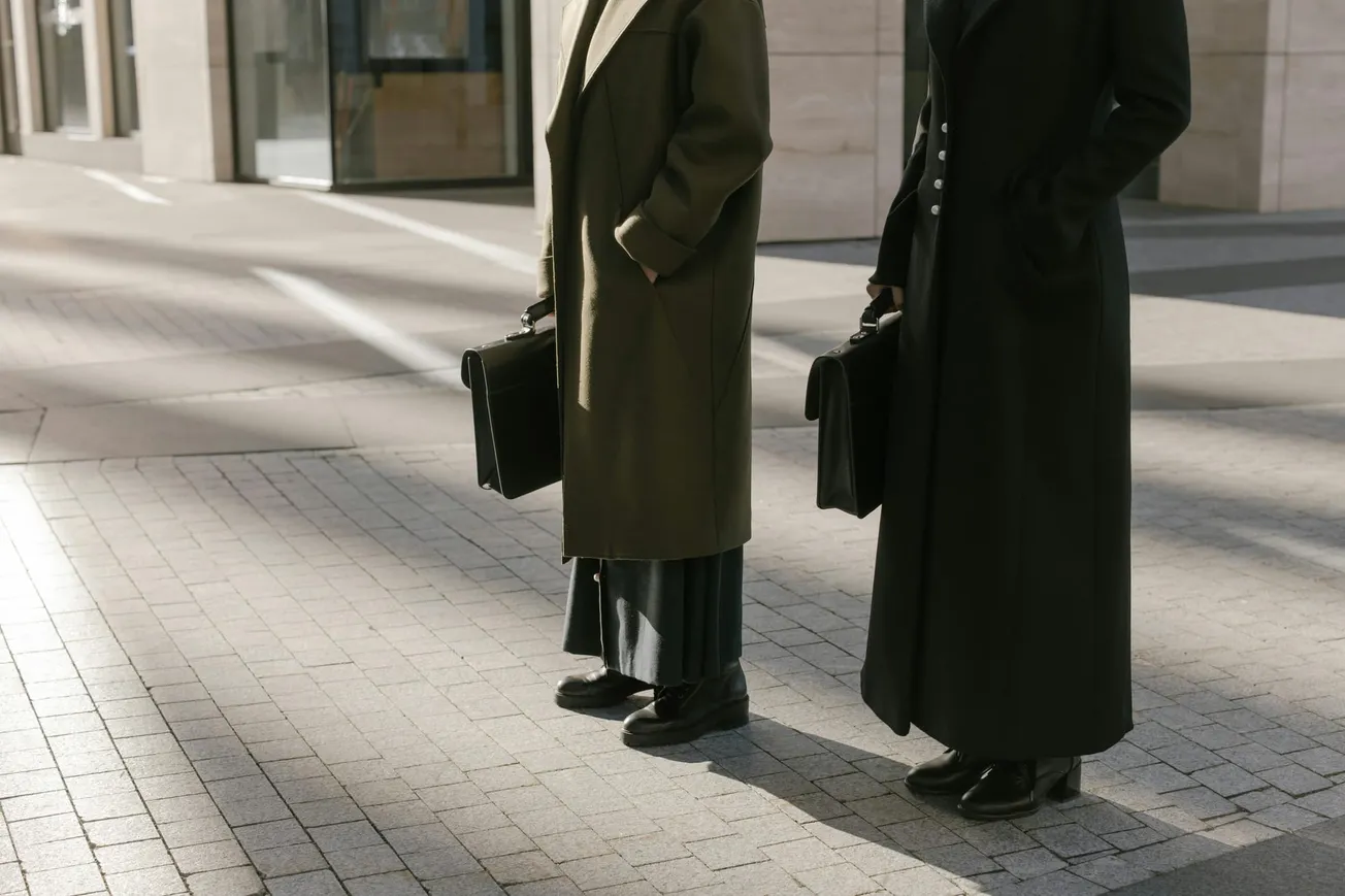 Two people in long coats stand on a sunlit, paved sidewalk holding briefcases. Their formal attire and posture suggest professionalism.