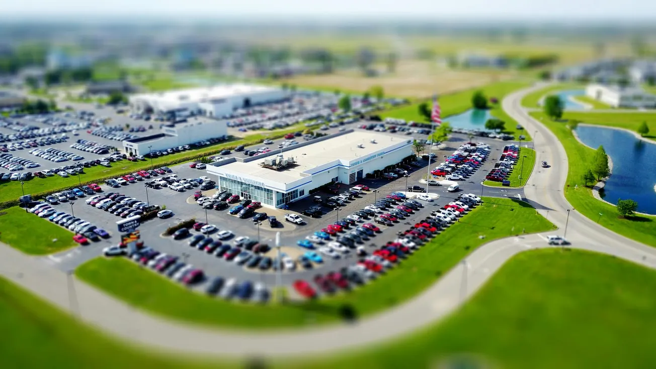 Aerial view of a large car dealership surrounded by numerous parked cars on a bright day. Curved roads and green lawns frame the scene, suggesting business and activity.