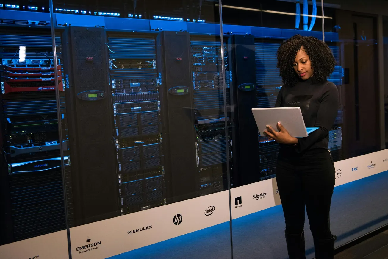 A woman stands with a laptop in front of illuminated server racks behind glass. The setting is clean and modern, conveying a tech-focused environment.
