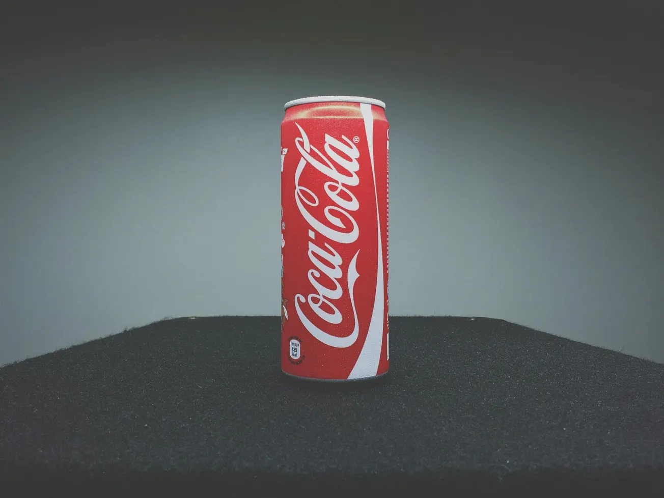 A red Coca-Cola can stands upright on a black surface. The can's white logo is prominent against the muted gray background, creating a classic, nostalgic feel.