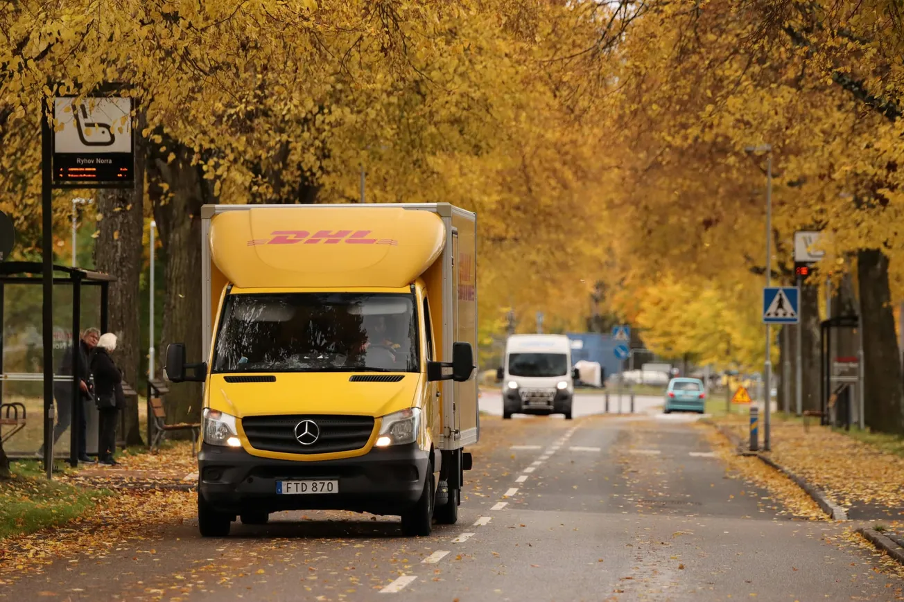 A yellow delivery truck drives down a tree-lined street in autumn. Golden leaves blanket the road, creating a warm and serene atmosphere.