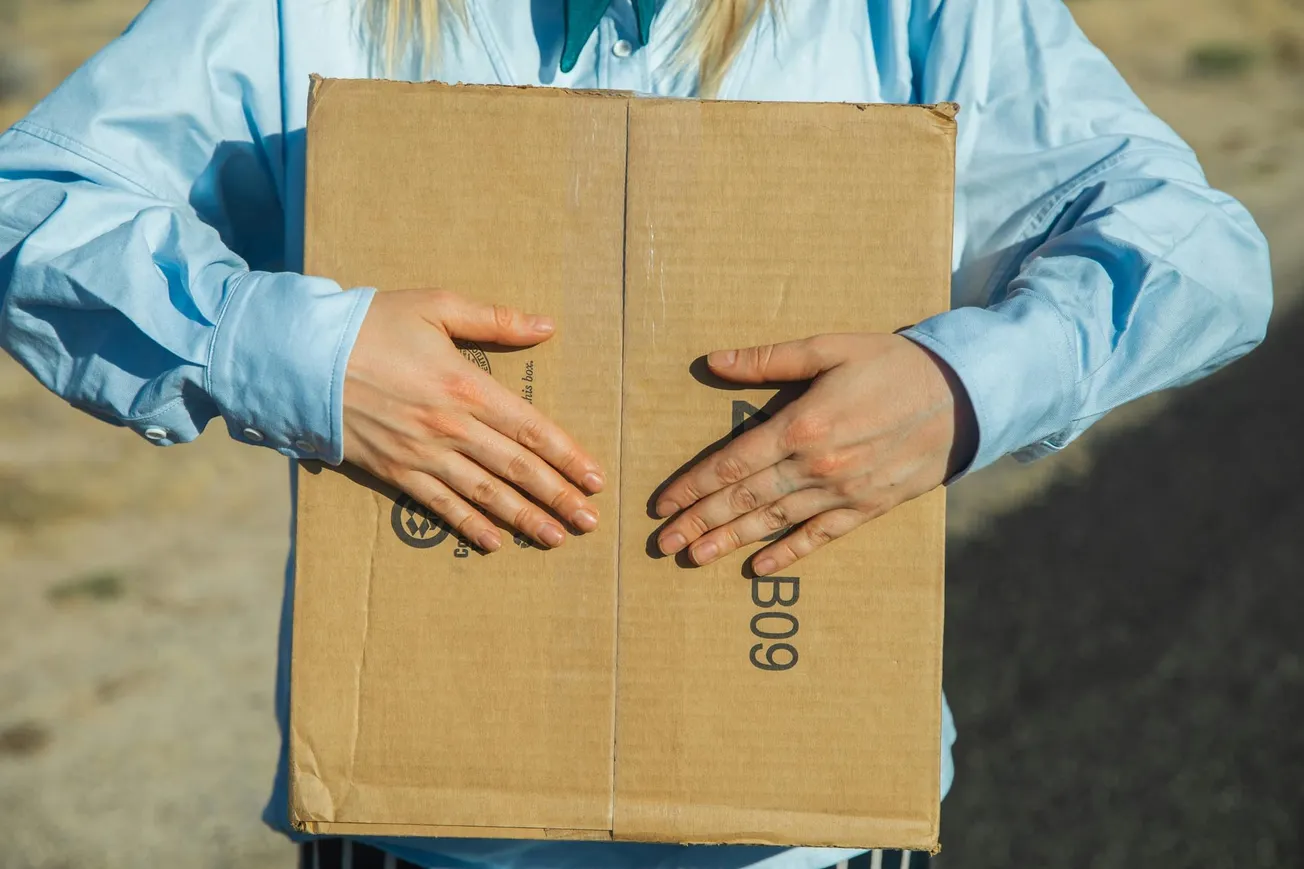 A person in a light blue shirt is holding a closed cardboard box. The setting is outdoors on a sunny day, with a blurred background of a road and dry grass.
