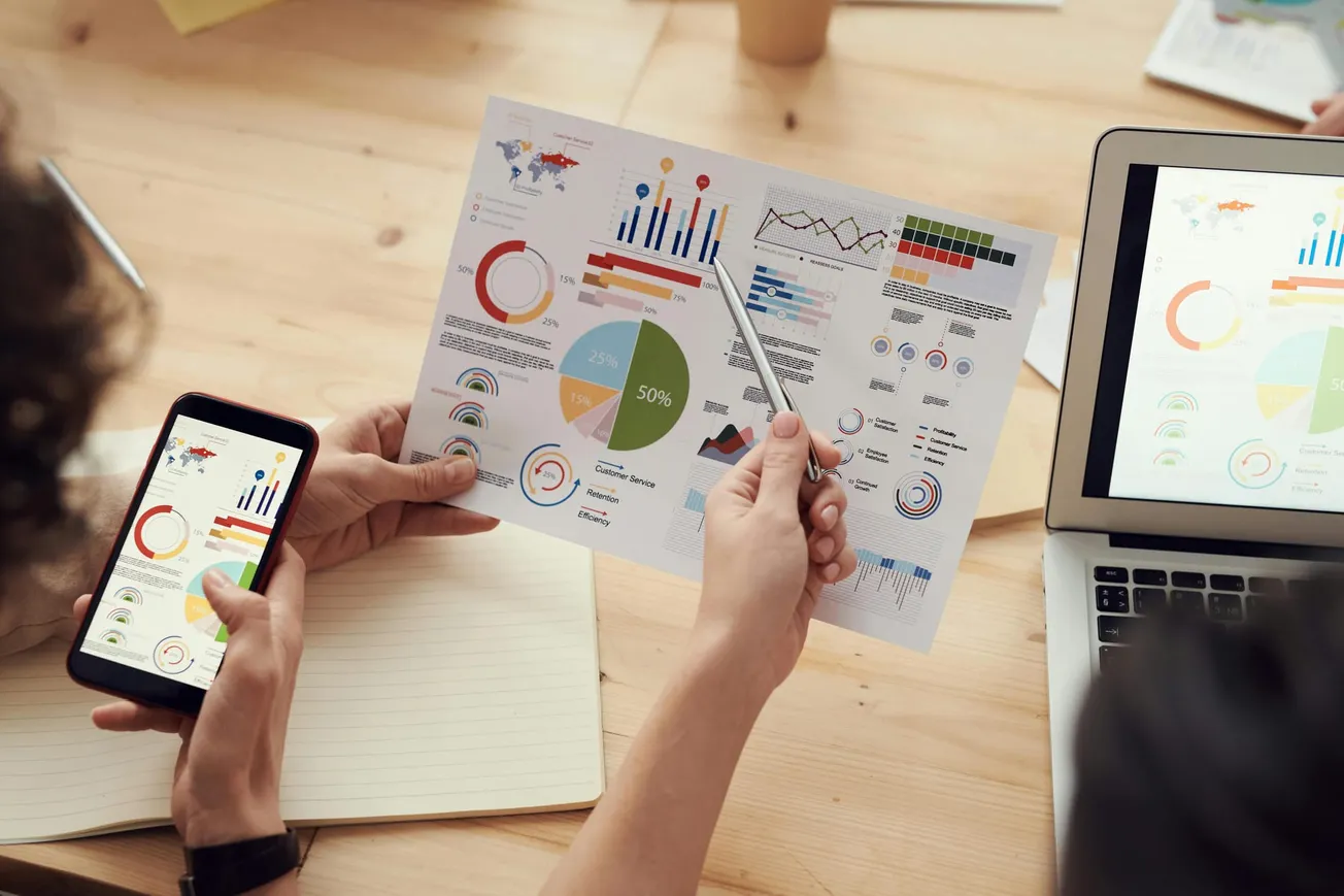 Two people examine colorful charts on a paper, smartphone, and laptop in a meeting, emphasizing data analysis and teamwork in a casual setting.