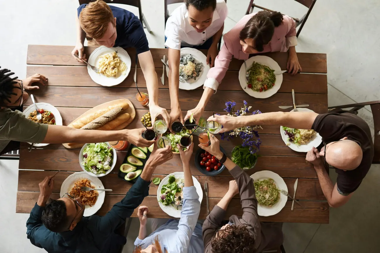 A diverse group of people sits around a wooden table enjoying a meal. They are raising glasses in a toast, surrounded by pasta, salad, bread, and flowers.