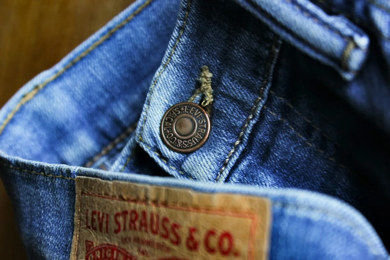 Close-up of blue jeans showing a brass button with "Levi Strauss & Co." engraved. A leather label in the foreground displays the brand name.