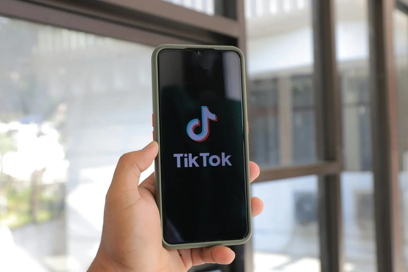 Hand holding a smartphone displaying the TikTok logo on a black screen. Background shows blurred windows, suggesting an indoor setting. Neutral tone.