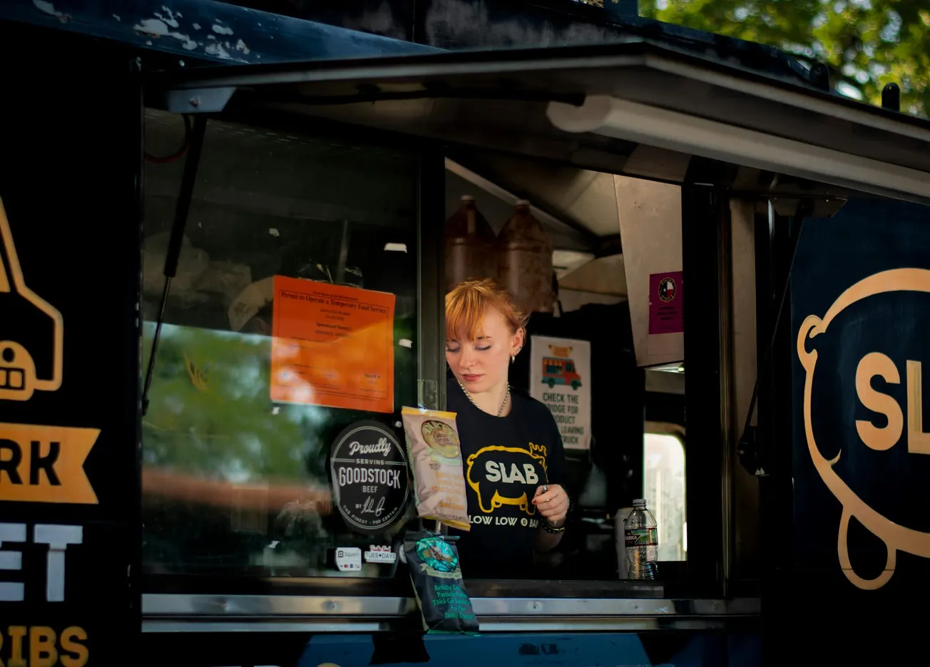 Young woman inside a food truck, reading a menu. She wears a "SLAB" shirt. The truck exterior has logos and colorful signs under sunny skies.