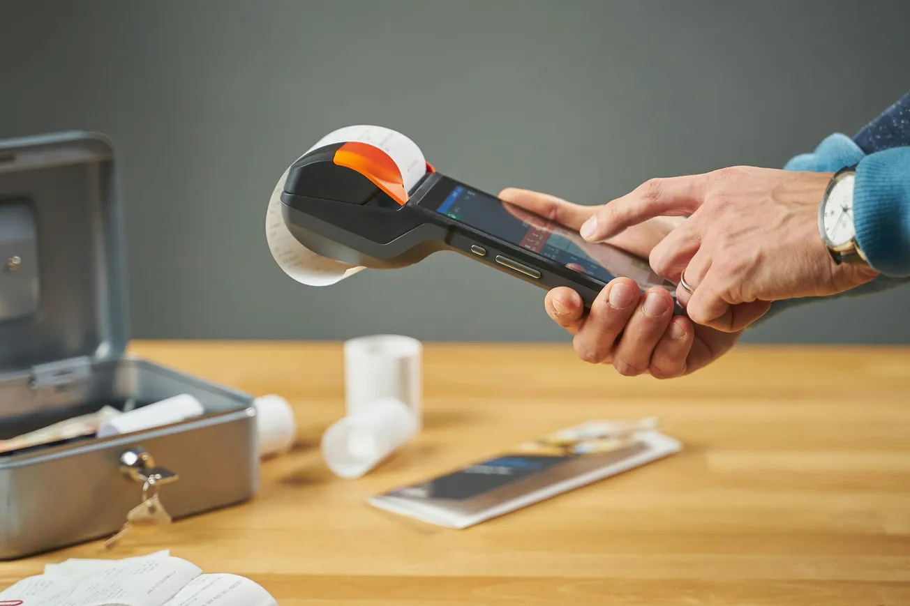 Hands holding a handheld receipt printer over a wooden table with receipts, a cash box, and paper rolls. The scene conveys efficiency and modern technology.