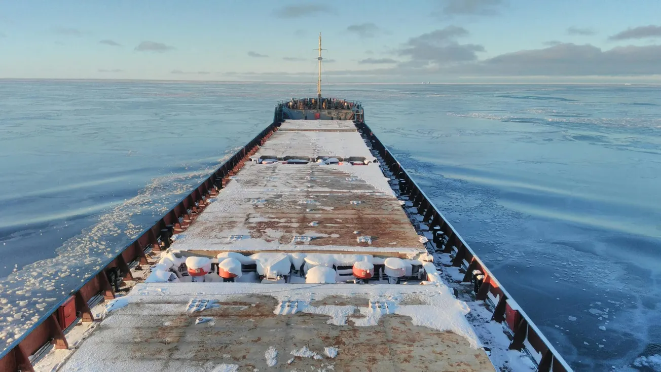 A large ship sails through icy waters under a clear blue sky. The deck is dusted with snow, contrasting with the rusty metal surface. Calm and serene.