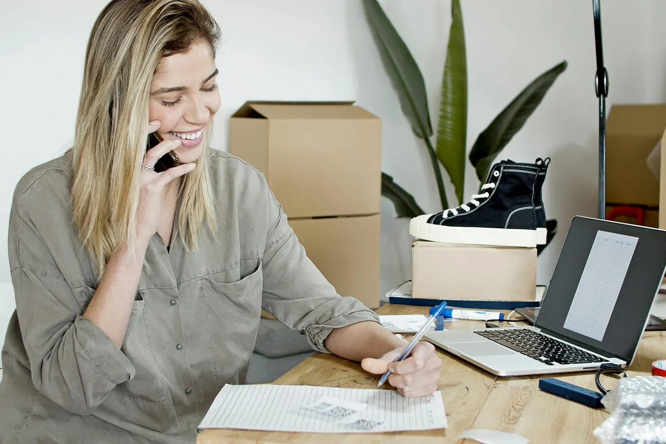 Smiling woman on phone writing notes at a desk with a laptop, shoe, and boxes. Bright, casual workspace.