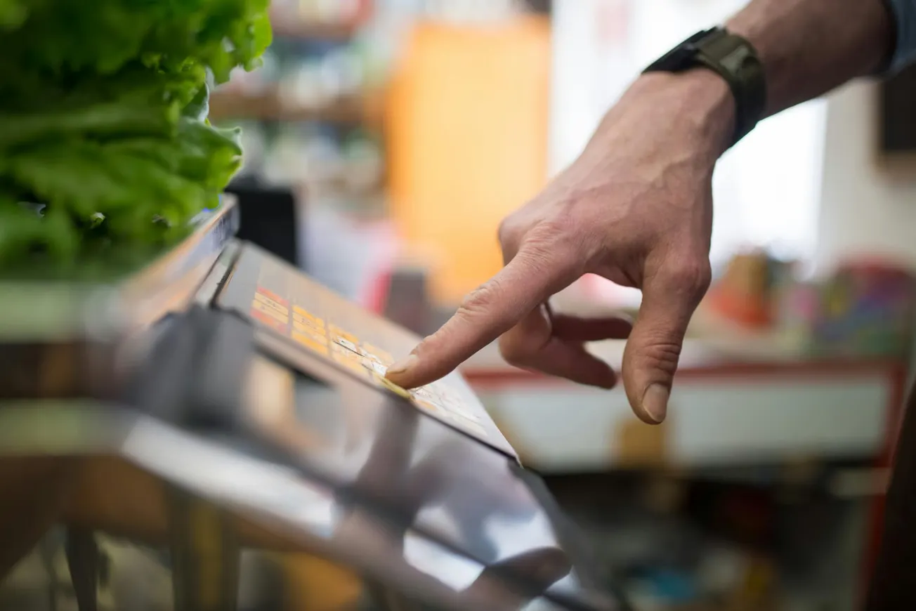 A hand with a watch presses a button on a digital scale next to fresh green lettuce. The scene suggests a grocery or market setting.
