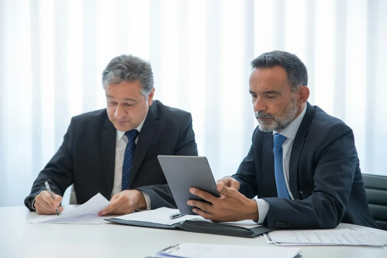 Two men in business suits sit at a table. One is writing on papers, while the other holds a tablet. They appear focused and serious.