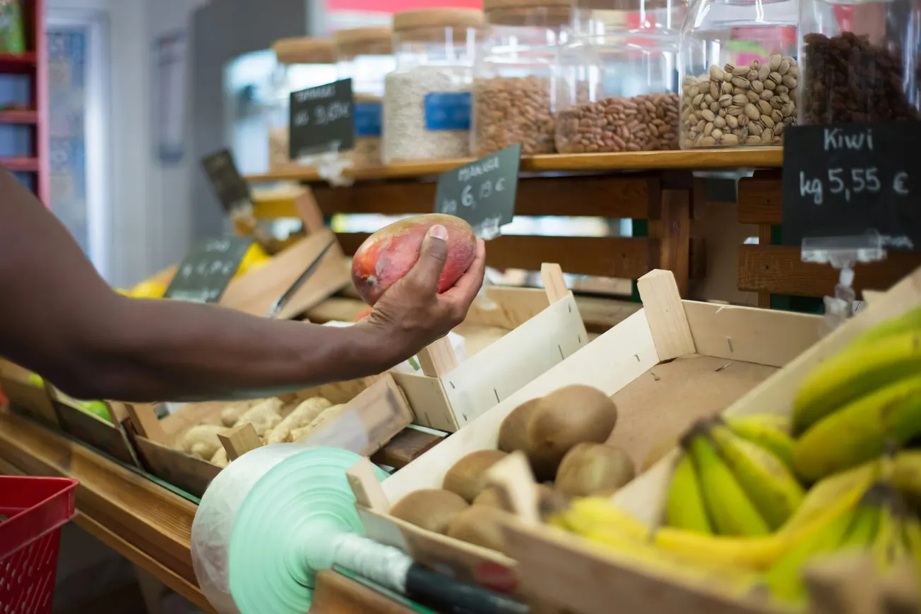 A person selecting a mango in a grocery store. The foreground shows bananas and kiwis, with jars of nuts in the background and price tags visible.