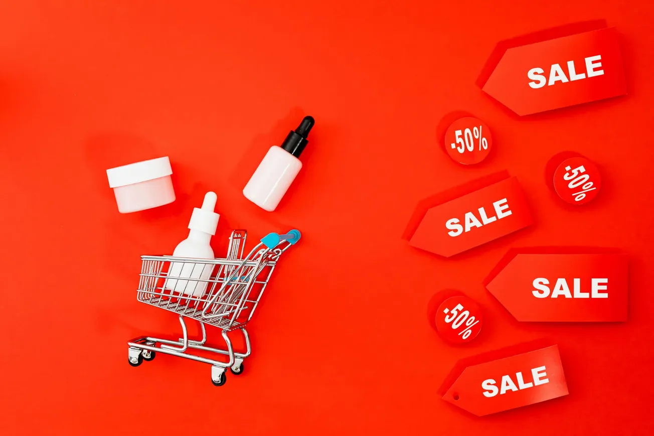 Shopping cart with skincare products on red background, surrounded by red sale tags reading "SALE" and "-50%," suggesting a discount promotion.