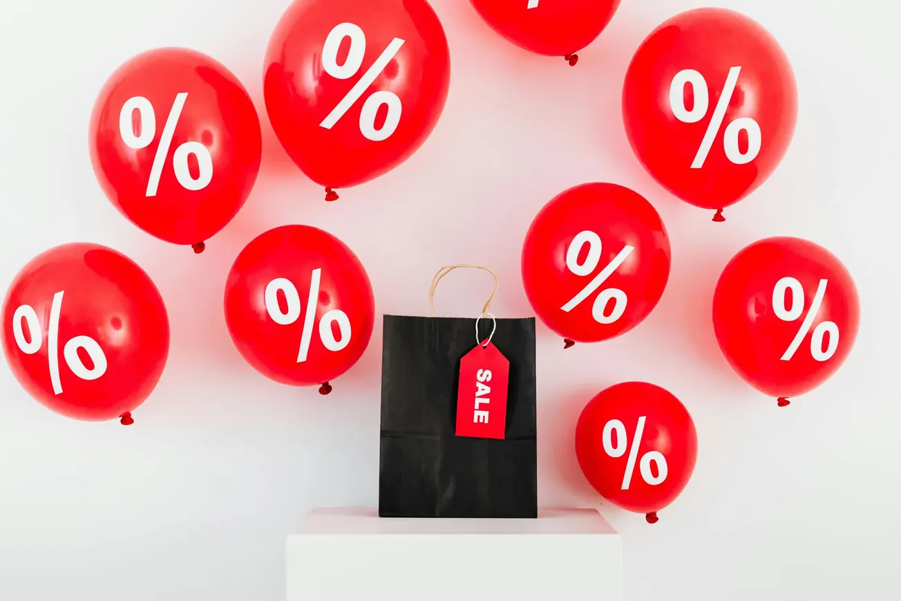 Red balloons with white percentage symbols surround a black shopping bag on a white pedestal. The bag has a red "SALE" tag, conveying a festive discount theme.