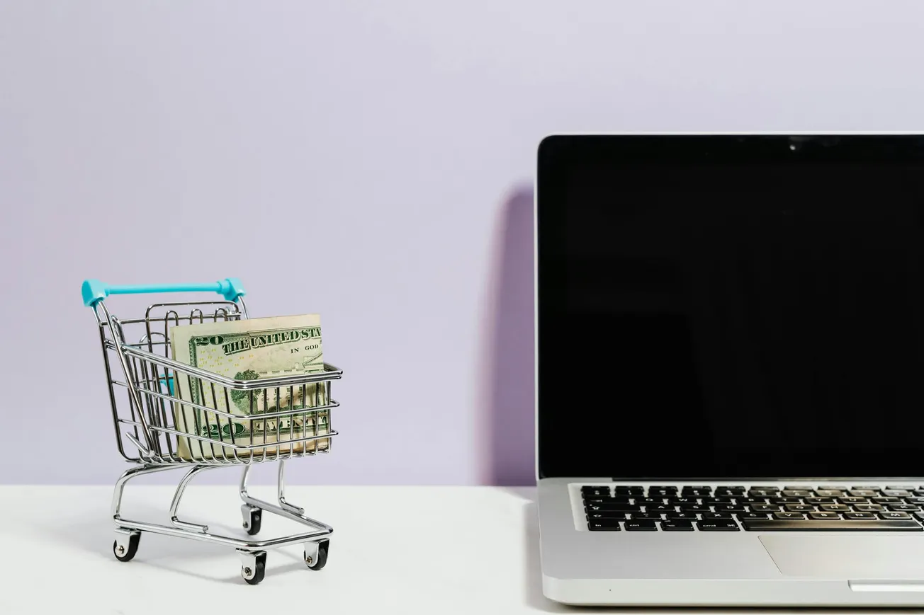 A small shopping cart filled with U.S. dollar bills is on the left beside a laptop with a blank screen on the right, set against a light purple background.