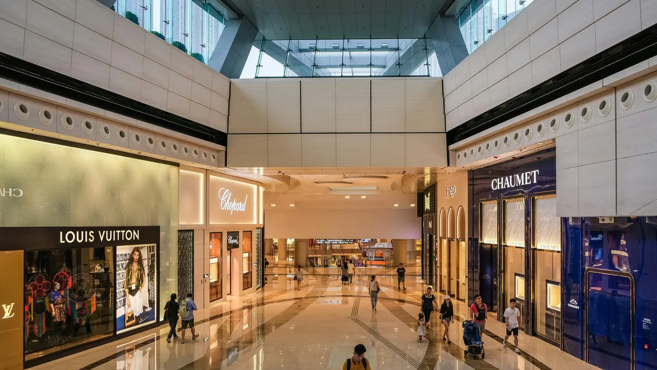 A modern, spacious shopping mall walkway with luxury stores like Louis Vuitton and Chaumet on both sides. Shoppers and bright, sleek design create an upscale vibe.
