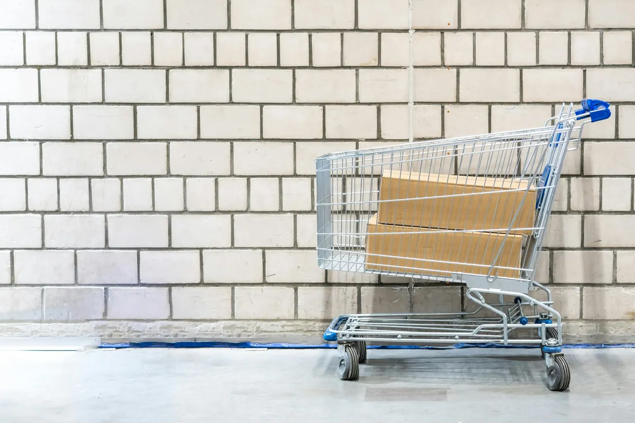 Shopping cart with cardboard boxes stands against a white brick wall on a concrete floor, evoking a sense of minimalism and simplicity.