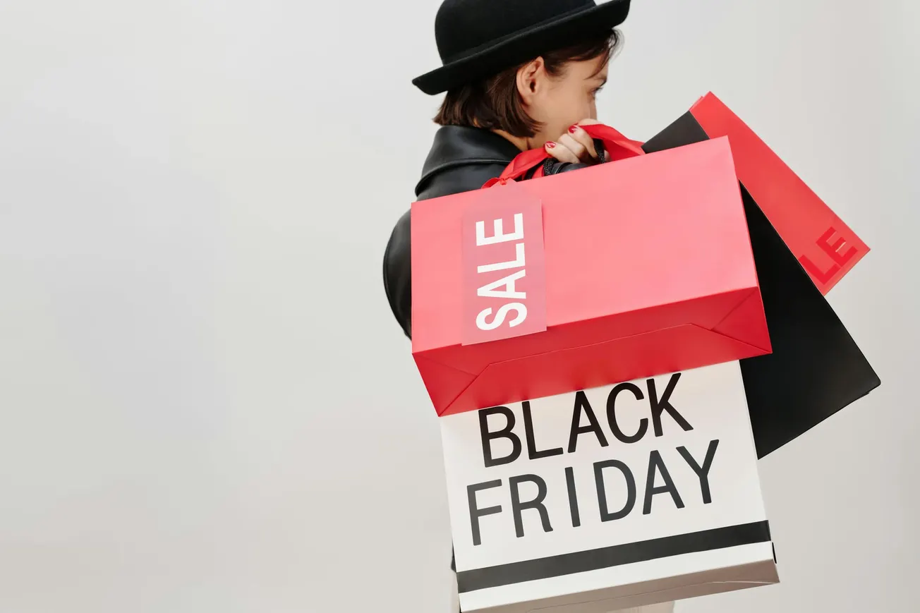 A person in a black hat holds several shopping bags. Two are red with "SALE" text, and one is white with "BLACK FRIDAY."