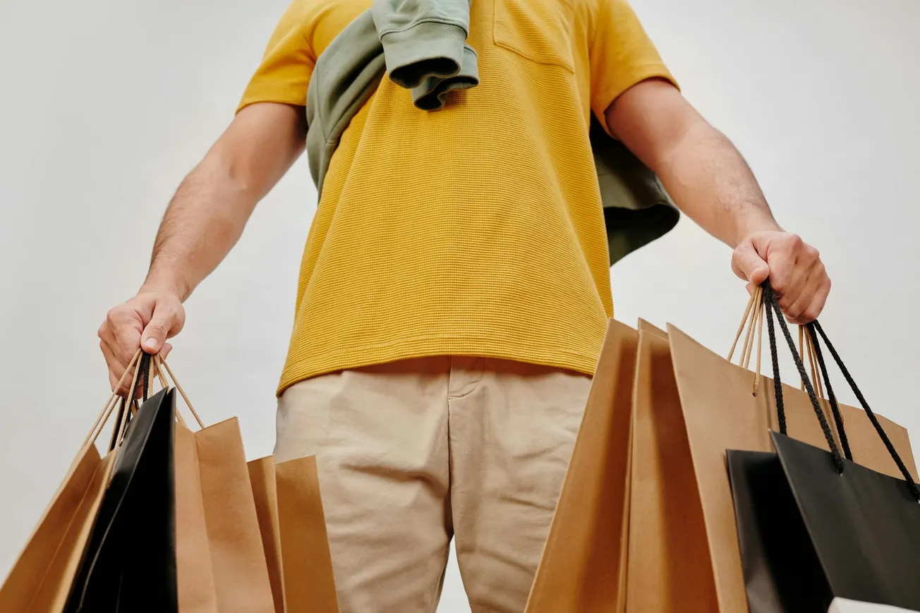 Man in a yellow shirt holds multiple brown and black shopping bags. A jacket is draped over his shoulder, conveying a casual shopping vibe.